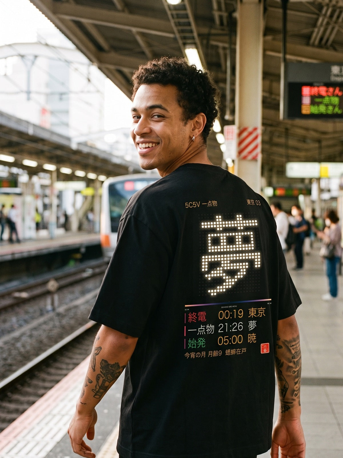Model on a Tokyo train platform looking back over his shoulder, wearing a black Asobi tee with a dot-matrix 夢 departure board on the back