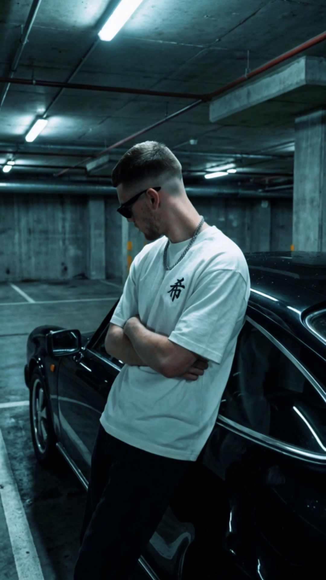 Man wearing a white 5C5V T-shirt with a small kanji chest print, leaning on a black car in an underground car park