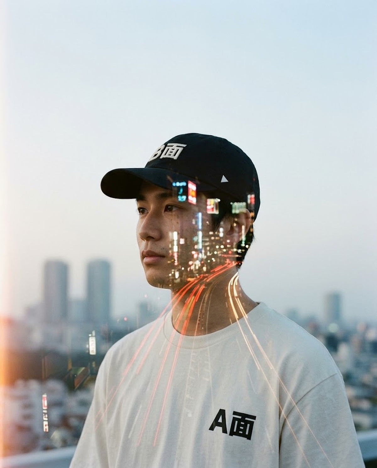 Double-exposure film photo: young man in a black B面 cap and cream A面 tee, Tokyo city lights over his face at dusk — 5C5V REWIND 巻き戻し