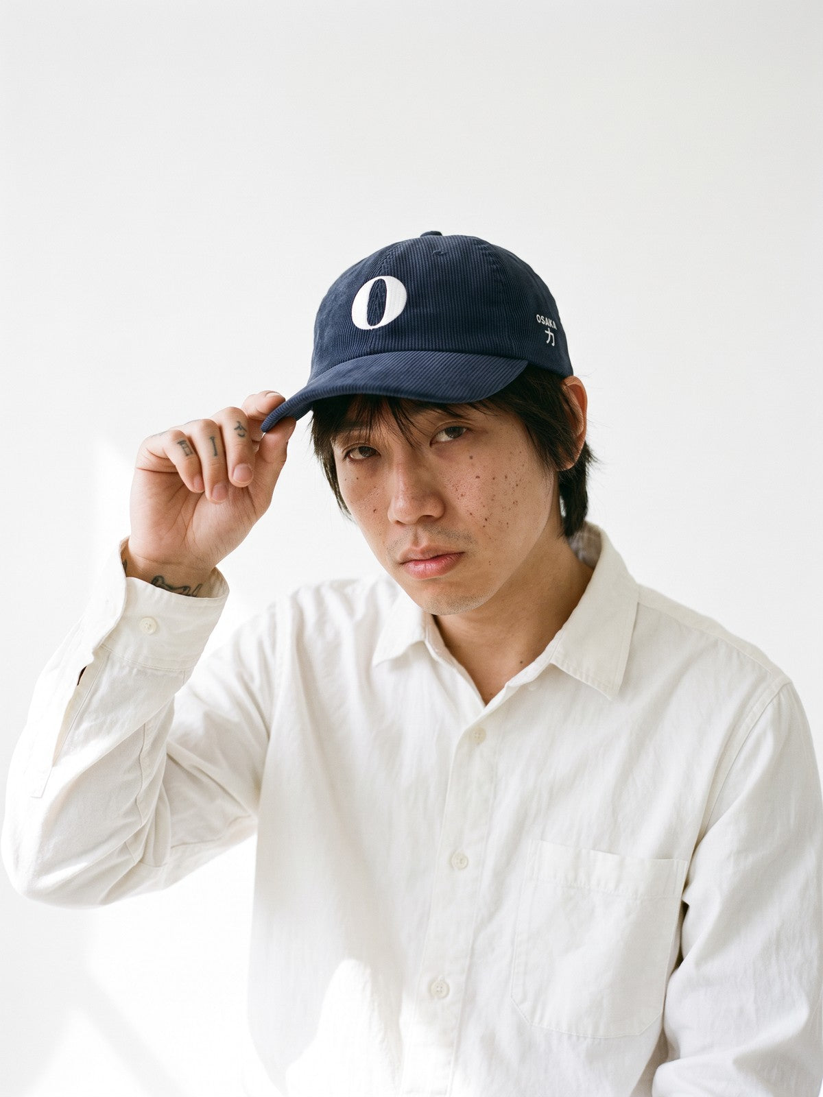 Model wearing the 5C5V Osaka O Corduroy Japanese baseball cap in navy, a serif O embroidered on the front, OSAKA and 力 (strength) on the left side