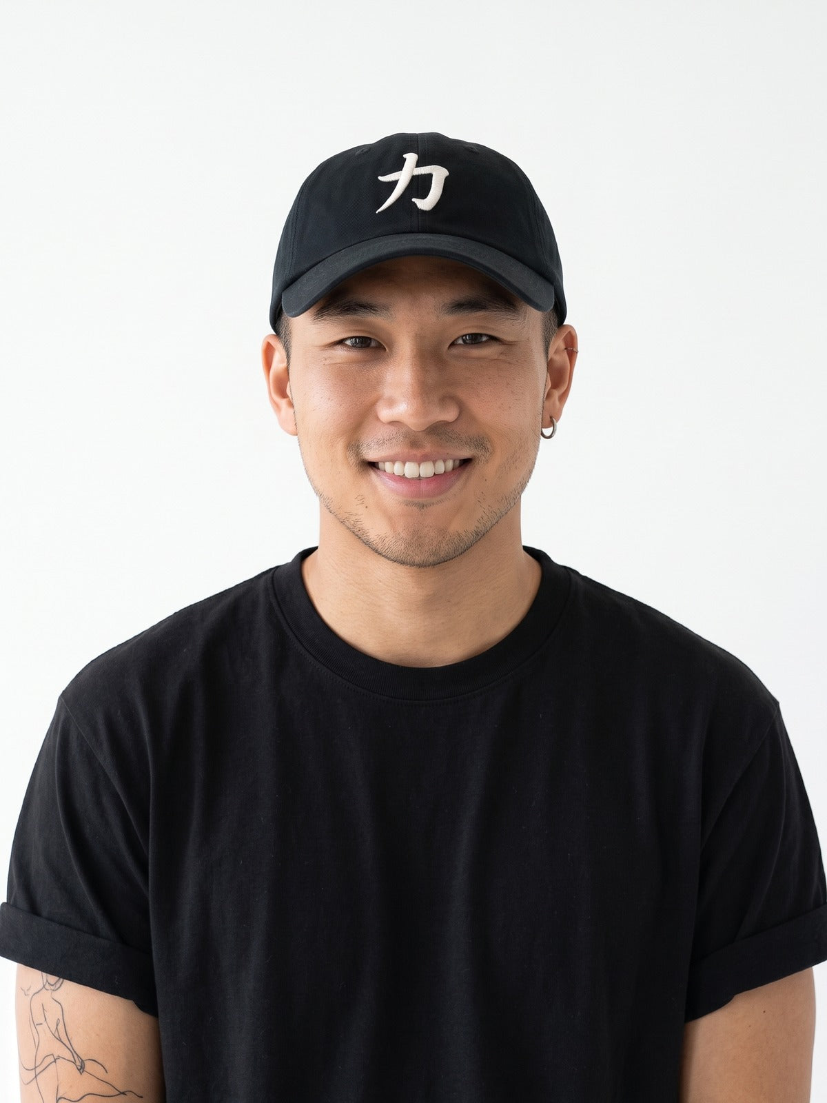 Smiling model with a silver hoop in a black tee wearing the black 5C5V Osaka Strength cap, white 力 (strength) embroidered on the front, close front view