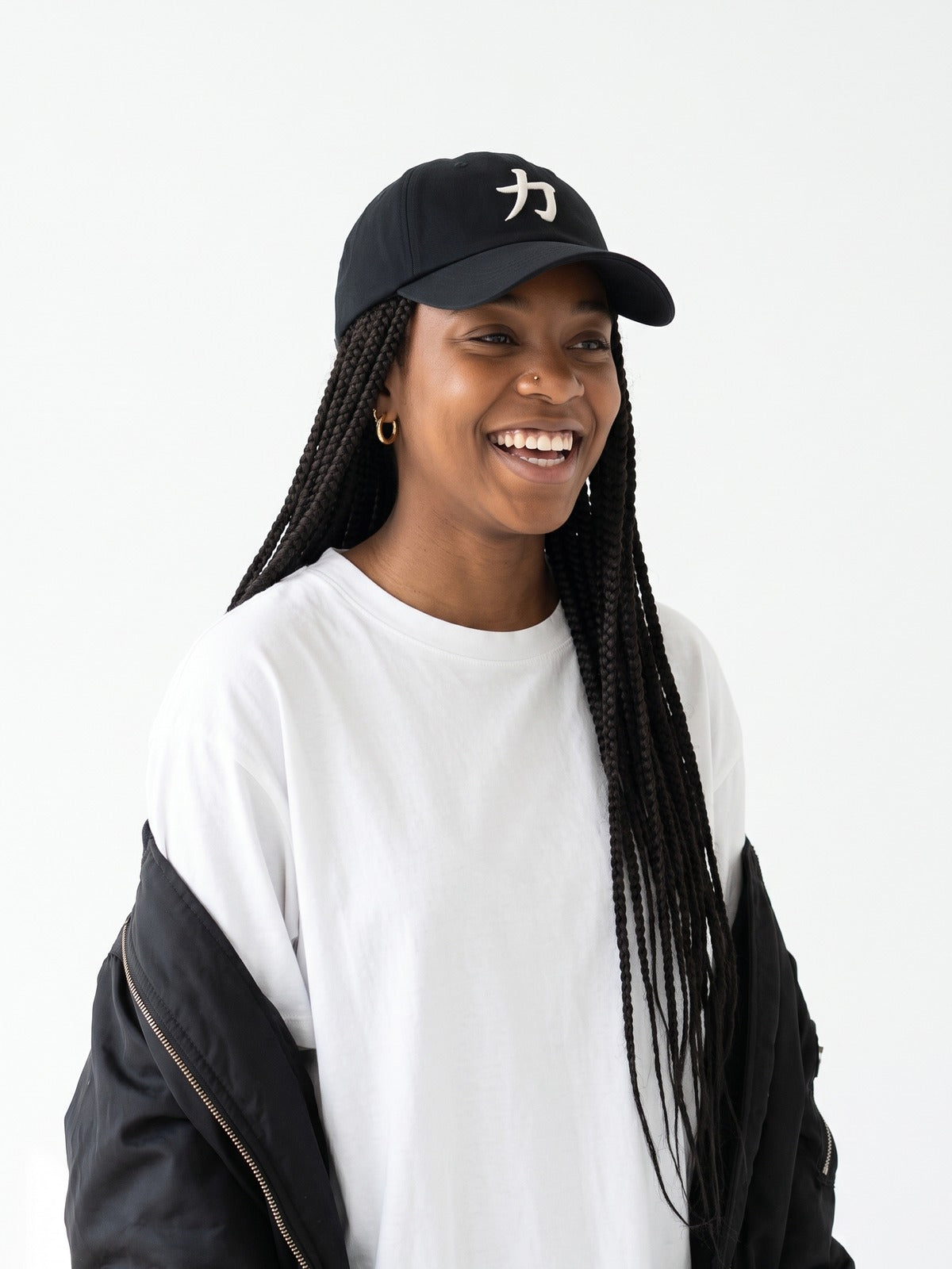 Laughing model with long box braids in a white tee and black bomber wearing the black 5C5V Osaka Strength 力 cap, three-quarter view