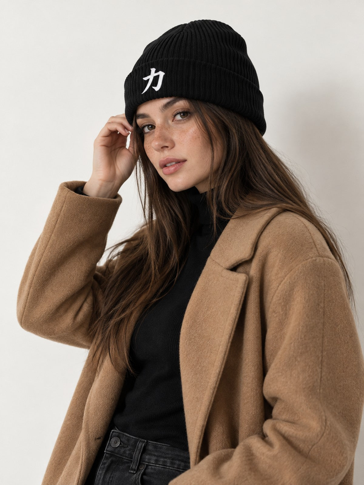 Model in a camel coat touching the cuff of the black 5C5V Osaka Strength 力 organic beanie, side angle