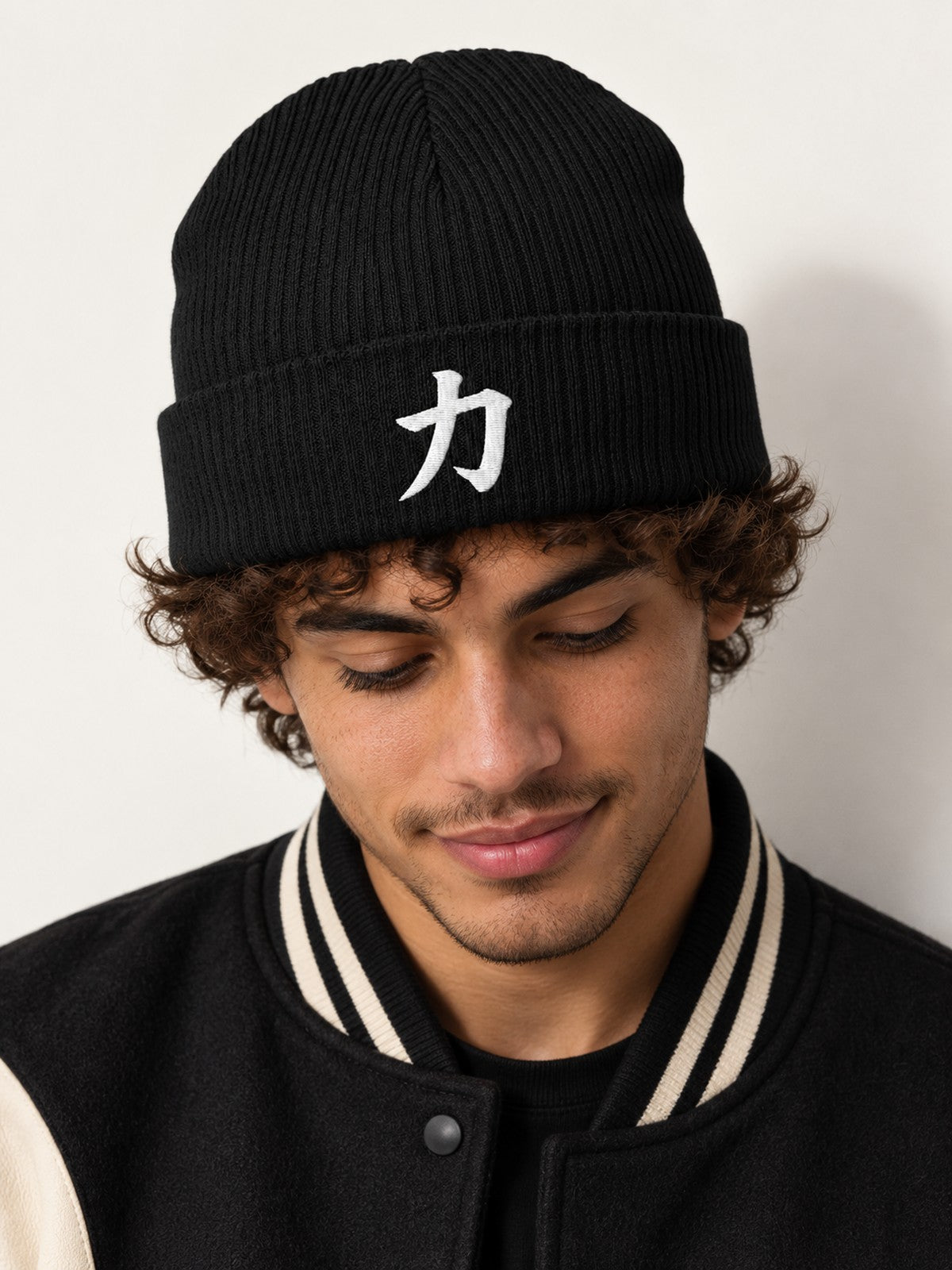 Close-up of the white embroidered kanji 力 (strength) on the black 5C5V Osaka organic beanie, worn by a model in a black and cream varsity jacket