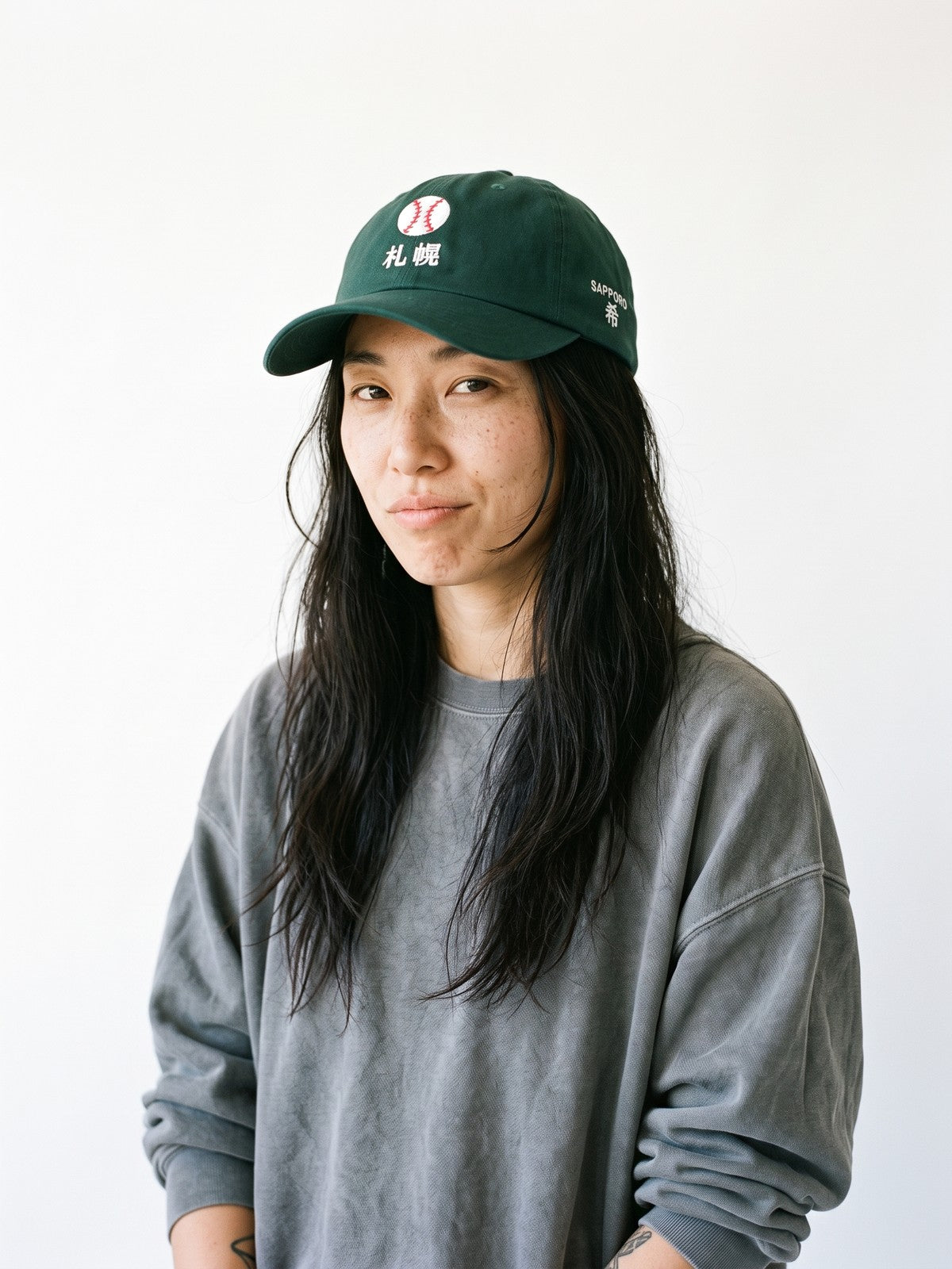 Model wearing the 5C5V Sapporo Ball Japanese baseball cap in spruce, a stitched baseball over 札幌 embroidered on the front, SAPPORO and 希 (hope) on the left side