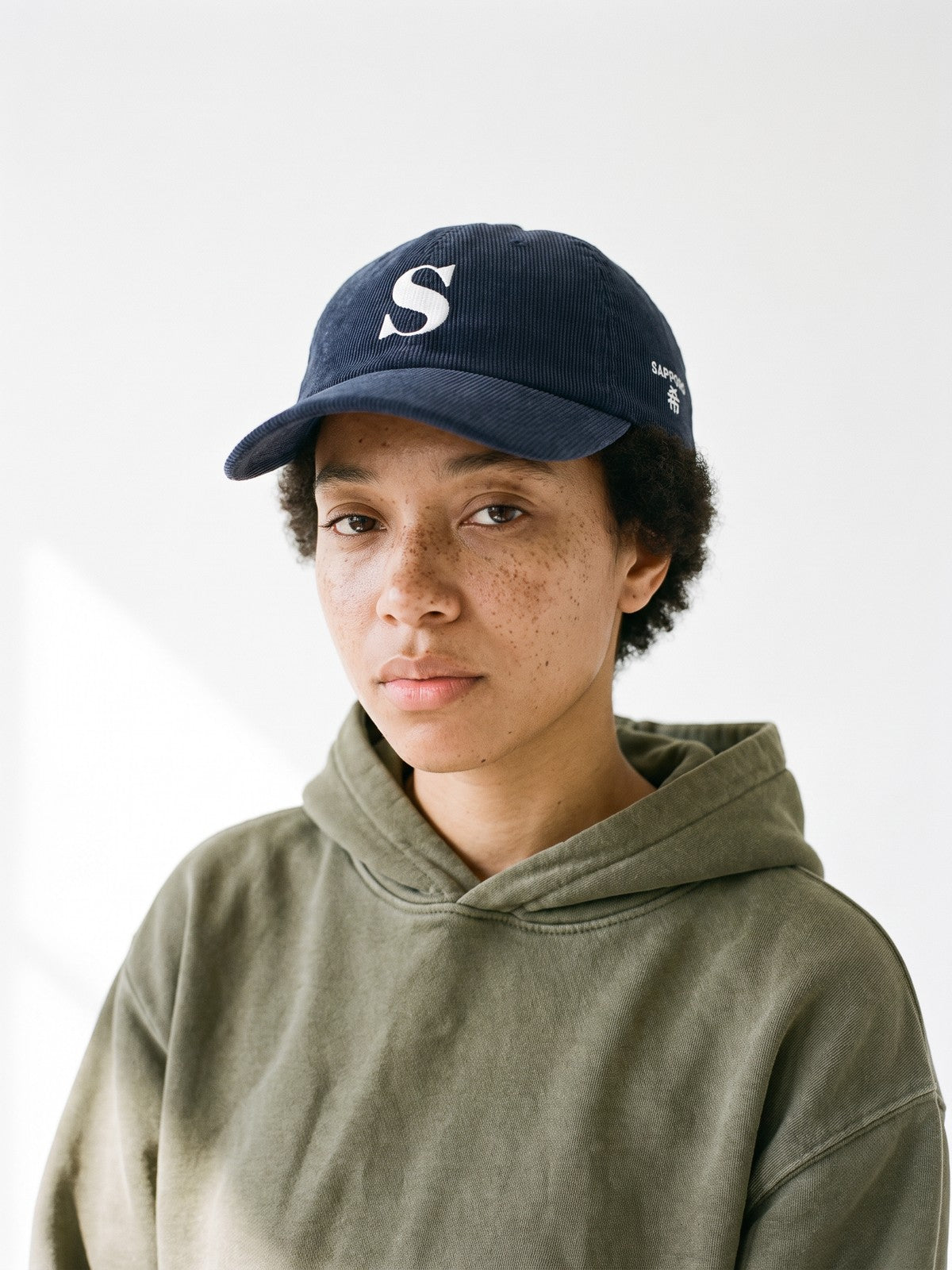 Model wearing the 5C5V Sapporo S Corduroy Japanese baseball cap in navy, a serif S embroidered on the front, SAPPORO and 希 (hope) on the left side