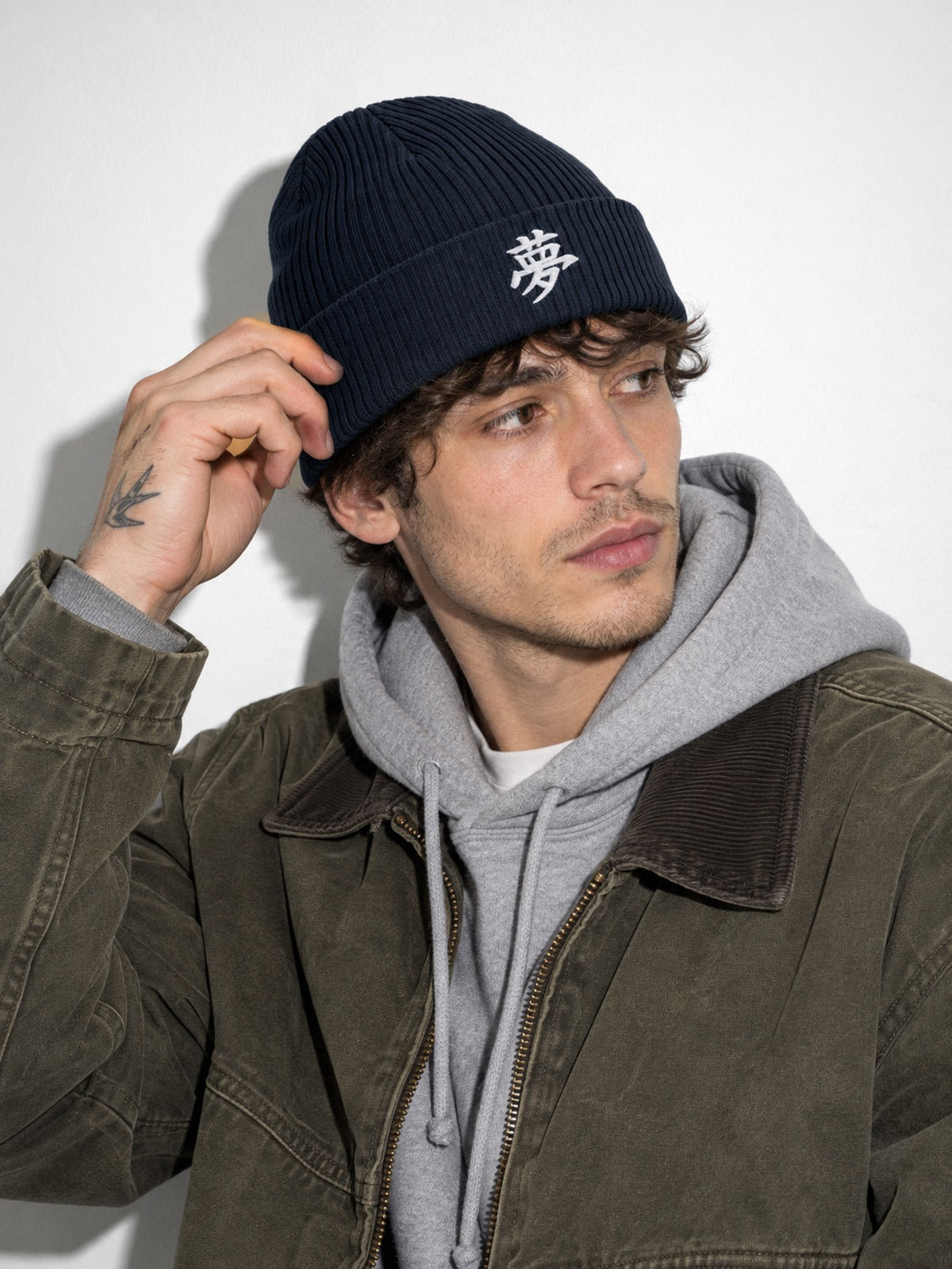 Model in an olive jacket and grey hoodie adjusting the cuff of the Navy 5C5V Tokyo Dream 夢 beanie, side angle
