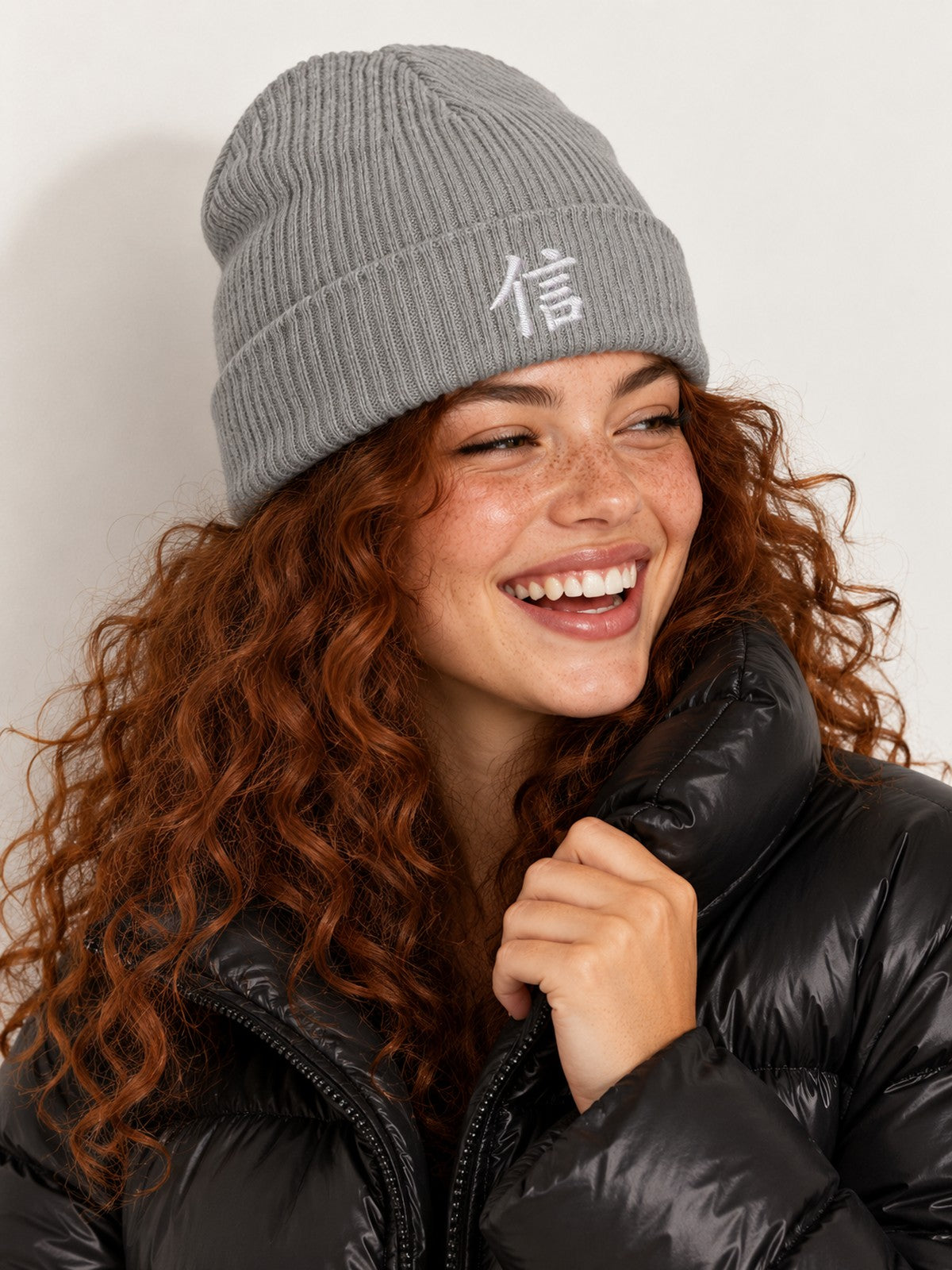 Laughing model with curly auburn hair in a black puffer wearing the Light Grey 5C5V Yokohama Trust 信 ribbed beanie, three-quarter view