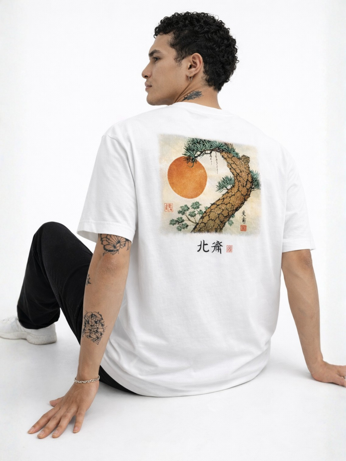 Seated model seen from behind wearing the white Seán Seachtain Hokusai tee, showing the large colour back print of a pine tree and rising sun — an Edo-period ukiyo-e woodblock homage signed with the 北斎 (Hokusai) seal.