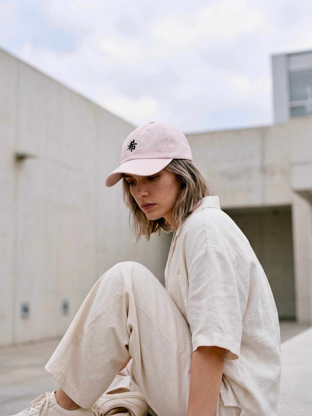 Model sitting against a concrete wall in the pastel pink First Light 希 cap and cream linen