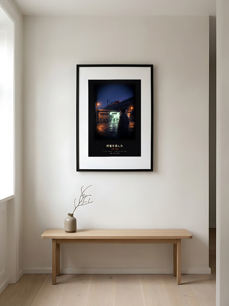 Shūden 終電 limited edition framed print: a young man in headphones outside a Tokyo station at midnight as the last train pulls away, black frame in a Japandi entryway above an oak bench