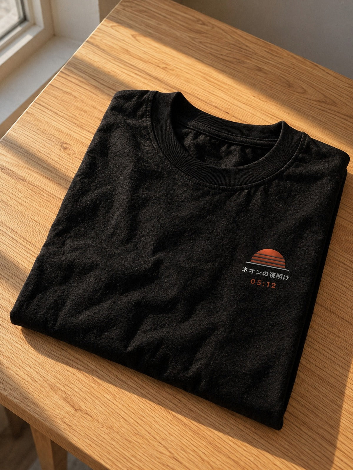 Neon Dawn Tokyo Bay 東京湾 heavyweight black tee folded on an oak table in late afternoon light, with the embroidered 05:12 sun on the chest